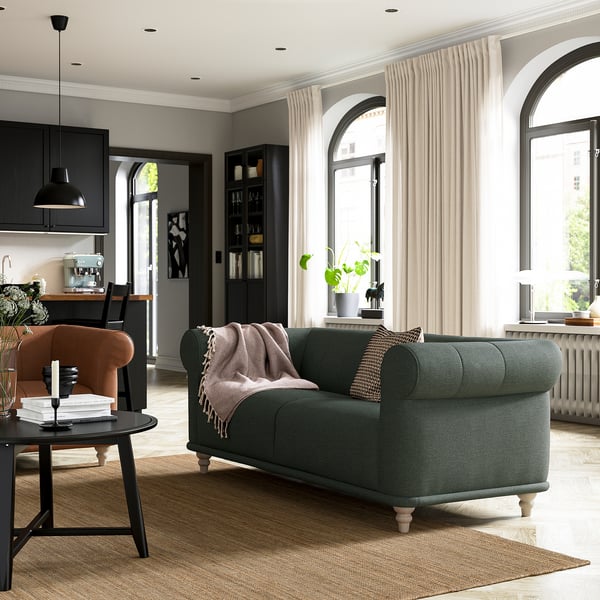 Moderno salón con sofá verde VISKAFORS, silla naranja, mesa de centro negra y grandes ventanales con cortinas.