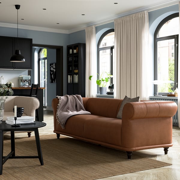 Sala de estar moderna con sofá VISKAFORS marrón, mesa de centro negra, grandes ventanas arqueadas y gabinetes de cocina oscuros.