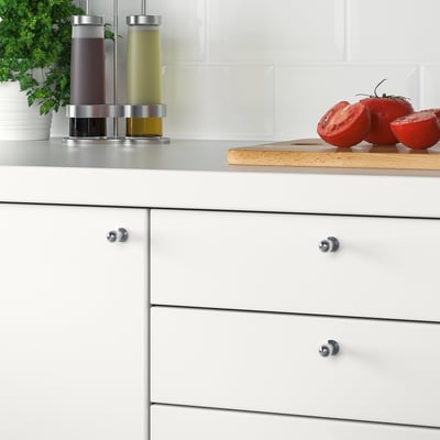 Sukaldeko armairu modernoak, BAGGANÄS pomo zilar-kolorekoekin, altzairu herdoilgaitzezko osagarriak eta tomate freskoak mozteko taula batean.