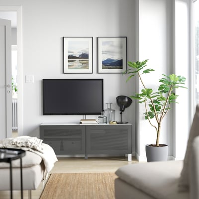 Telebista beltzeko gela modernoa, BESTÅ euskarri grisean, naturako bi estanpatu, landarea eta sofa zuria.
