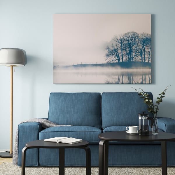 Sofa urdina, pastel-paisaia, zutik dagoen lanpara, dekoraziodun mahai osagarriak.