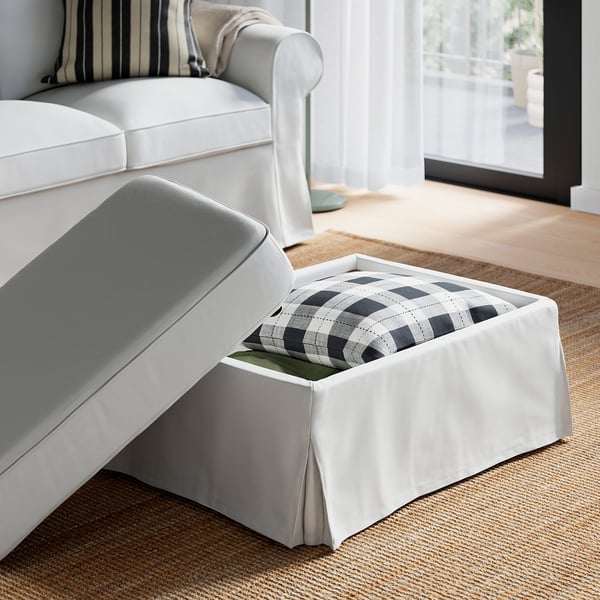 Sofa zuria, ottomanarekin, alde bat zabalik, barruan zuri-beltzezko manta xakeatua erakusten duena, leiho handi baten ondoan alfombra testuradun baten gainean jarrita.