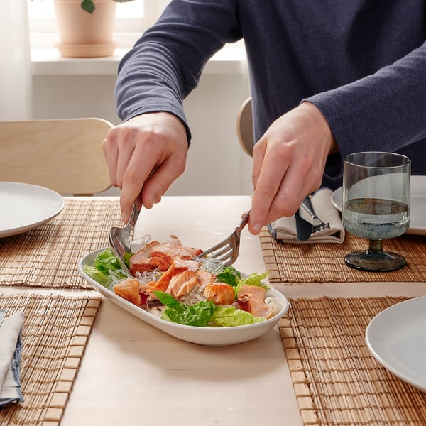 Pertsona bat entsalada jaten sardexka eta katilu zuri baten koilara mahai gainean, edalontzi eta platerekin.