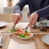 Pertsona bat entsalada jaten sardexka eta katilu zuri baten koilara mahai gainean, edalontzi eta platerekin.