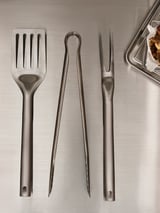 Altzairu herdoilgaitzezko erretegi hiru erreminta: pintzeak, espatula eta sardengailua, metalezko gainazal baten gainean jarrita. Diseinu dotorea, parrilla erretzeko praktikoa.
