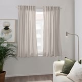 HANNAILL gortinak, beige argi kolorekoak, kotoizkoak, zoruraino, sinpleak eta dotoreak, landare handi baten eta sofa zuri atsegin baten ondoan zintzilik daude, kukor berdeekin.