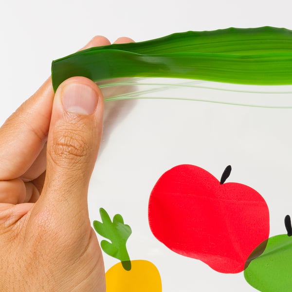 Eskua hosto berde fresko bat eskuan, atzeko planoan fruta eta barazki koloretsuen 3D erreprodukzioekin.