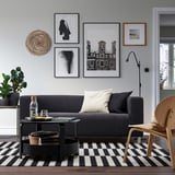 Egongela modernoa, KLIPPAN sofa grisarekin, erdiko mahai dotorea, zuri-beltzeko koadrodun alfonbra, arte galeriako horma eta egurrezko aulkiak.