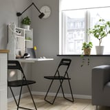 Bulegoko konfigurazio minimalista modernoa, hormaren gainean mahai abailagarri grisa, aulki tolesgarri beltza eta egurrezko zoru gaineko lorontzietan landareak dituena.