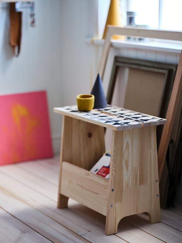Egurrezko taburete argia, azulejo zuri eta beltzen estalkia, kikara hori txikia, kono urdina, arte-lan koloretsuak, argi naturala, diseinu minimalista.