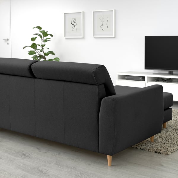 Sofa sekzional gris ilun modernoa, bizkarralde altua, lerro garbiak eta horma zuriaren kontra egurrezko hankak dituena, landareak, telebista eta argazkiak lagun dituela.