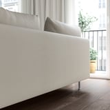 Sofa modernoa, gris argia, eserleku sakonak, hanka altuak, leiho handi baten ondoan.