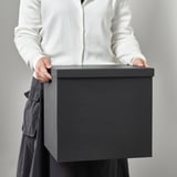 Pertsona bat, TJENA kaxa angeluzuzen gris handi bati eusten; Multimedia gailuak edo denbora-pasako artikuluak biltegiratzeko egokia.