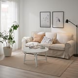 VODSKOV beige-ko jute-sisalezko alfonbra, sofa argi baten azpian, erdi-zuriko mahai txiki bat eta leiho handi bat.