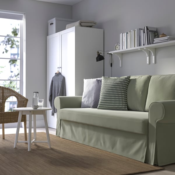 Sofa-ohea VRETSTORP berdea egongelan, armairuaren ondoan, liburuen apalaren azpian. Diseinu minimalista, egurrezko zoruak, mahaia eta aulki zuria dituena.