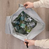 Unha persoa que sostén unha bolsa de reciclaxe reutilizable aberta chea de latas e botellas baleiras, mostrando o seu uso para a clasificación de residuos.