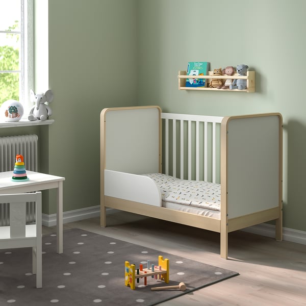 Gardería moderna con cama de bebé ÄLSKVÄRD de madeira clara, alfombra branca, apilador de xoguetes e estante creando espazo acolledor.