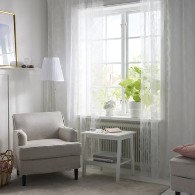 Salón luminoso e aireado con cortinas de encaixe brancas ALVINE SPETS , butaca gris, pequena mesa branca con plantas e libros, lámpada e cesta de vimbio.