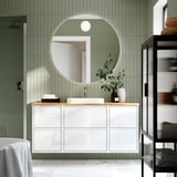 Baño moderno con vaidade branca ÄNGSJÖN, espello redondo, paredes verdes, chan de mármore e estante negro. O lavabo e a mesa son centrais e amplos.