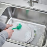 Persoa fregando o prato con cepillo verde en lavabo de aceiro inoxidable.