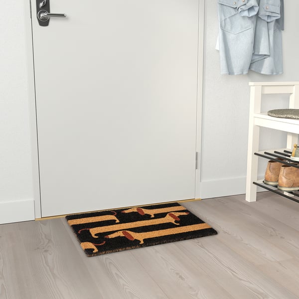 A alfombra de porta con deseño de dachshund negro e marrón atópase preto dunha porta branca e un estante de zapatos.