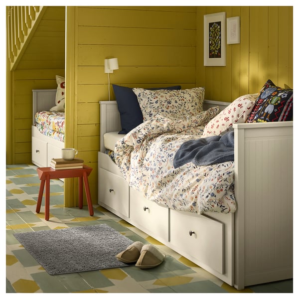 Dormitorio acolledor con paredes amarelas, camas brancas, roupa de cama floreada, un taburete vermello, alfombra gris e zapatillas.