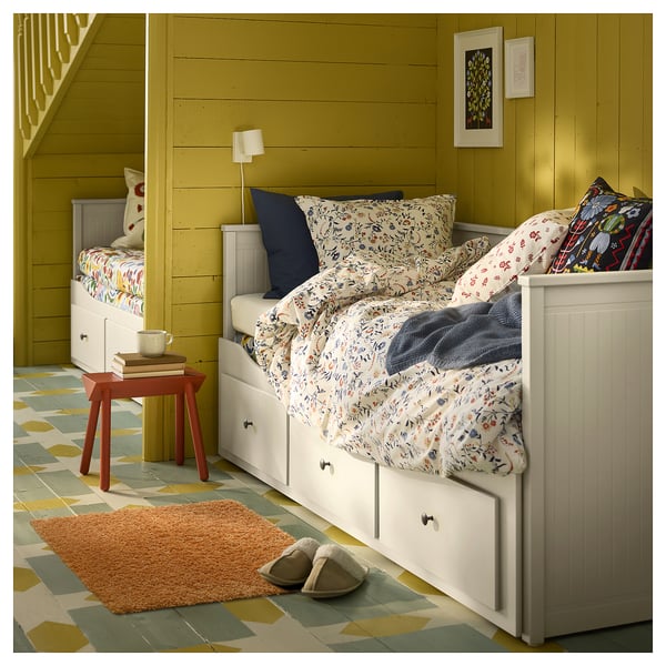 Un dormitorio acolledor con paredes verde oliva, camas brancas, roupa de cama floral, unha mesiña de madeira e unha alfombra marrón suave sobre un chan de azulexos xeométricos de cores.