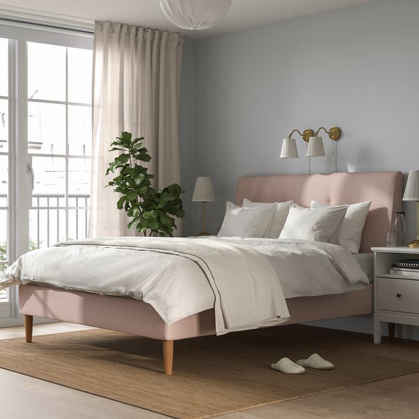 Imaxe do dormitorio con lámpadas rosas ÅRSTID na parede, cama grande con sabas brancas, planta por fiestra e zapatillas no chan.