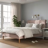 Imaxe do dormitorio con lámpadas rosas ÅRSTID na parede, cama grande con sabas brancas, planta por fiestra e zapatillas no chan.