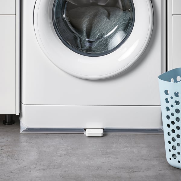 O pequeno sensor de fugas branco de BADRING está preto da lavadora, xunto á cesta da lavandaría azul, nun chan gris.