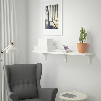 Unha habitación minimalista cunha cadeira de brazos gris, estante branco BERGSHULT montado na parede con libros, unha pequena planta de cactus e unha imaxe do bosque enmarcada.
