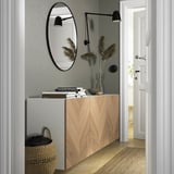 Entrada moderna con armario BESTÅ, madeira clara, espello redondo, luces de parede, plantas secas, libros e cesta de vimbio.