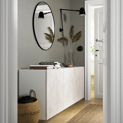 Armario branco BESTÅ con estantes e portas, montado na parede, flanqueado por lámpadas negras, libros e decoración con espello e cesta.