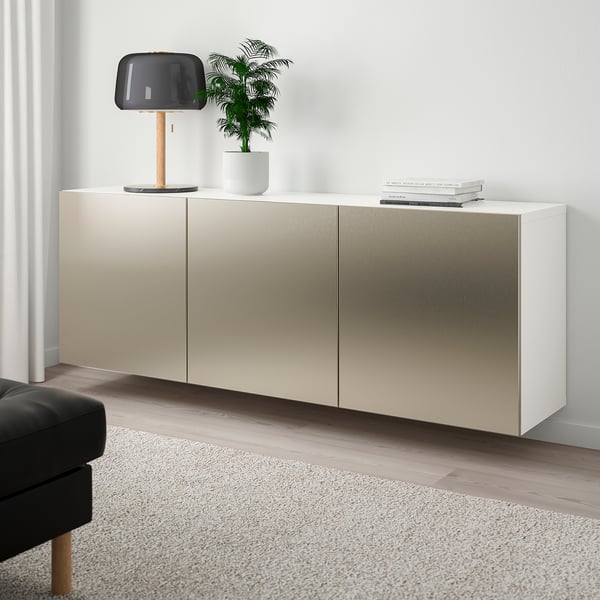Armario BESTÅ montado na parede en beige, con lámpada, planta e libros, con ottoman negro sobre alfombra gris.