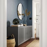 Moderno taboleiro gris BESTÅ con portas de malla, deseñado cunha lámpada, libros e vaso. Contra a parede azul escura, xunto á porta branca.
