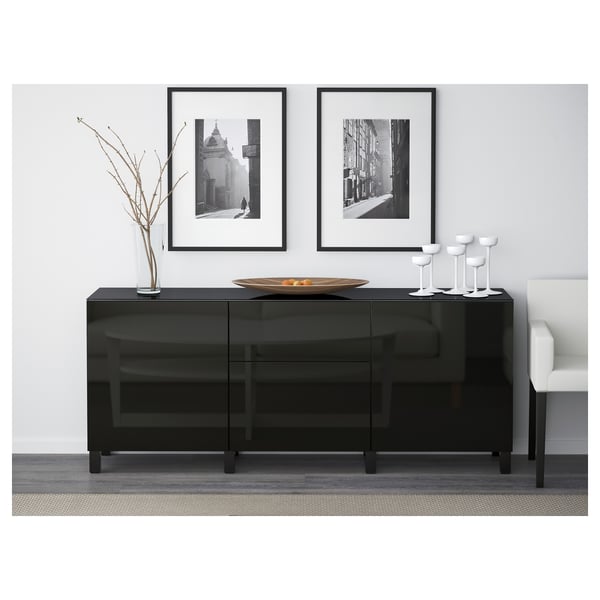 Moderno sideboard de vidro negro con caixóns e estantes. Rematada con cunca decorativa e vasos, contra unha parede branca con fotos enmarcadas.
