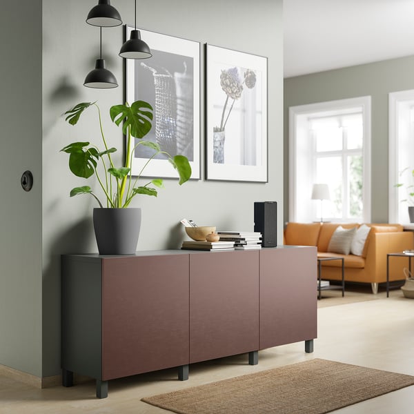 Sala de estar moderna con sideboard BESTÅ en marrón rosado; gran planta verde na parte superior; Sofá laranxa e luz natural desde a fiestra.