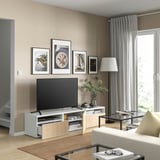Sala de estar moderna con banco de televisión BESTÅ en branco e carballo. Televisión de pantalla plana, arte enmarcada, vaso con verde e cortinas grises.