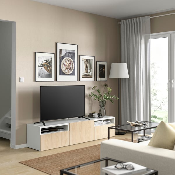 Sala de estar moderna con paredes beige, banco de televisión branco BESTÅ, televisión, marcos decorativos, sofá beige, mesa de café de vidro e xanela con cortinas grises.