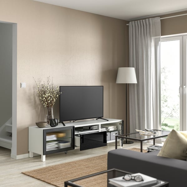 Sala de estar moderna con banco de televisión branco BESTÅ, televisión de pantalla plana, mesa de café con vidro, lámpada e gran xanela con cortinas.