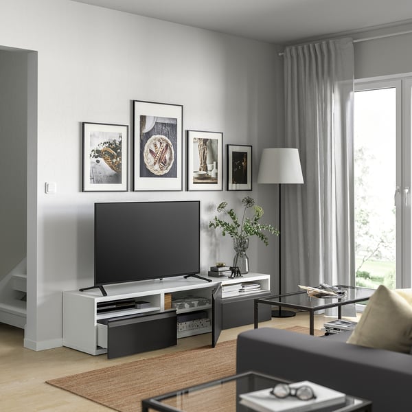 Sala de estar moderna con banco de televisión BESTÅ, televisión de pantalla plana, imaxes enmarcadas, lámpada de mesa, mesa de café de vidro, alfombra beige, cortinas grises.