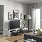 Sala de estar moderna con banco de televisión BESTÅ, televisión de pantalla plana, imaxes enmarcadas, lámpada de mesa, mesa de café de vidro, alfombra beige, cortinas grises.