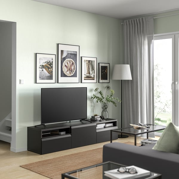 Sala de estar moderna cun banco de televisión BESTÅ negro, unha gran televisión de pantalla plana, fotos enmarcadas, unha cortina gris e unha mesa de café de cristal.