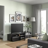 Sala de estar moderna con paredes verdes claras, banco de televisión BESTÅ negro, televisión de pantalla plana, arte enmarcada, cortinas grises, mesa de café de vidro, alfombra e planta en pota.