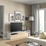 Sala de estar moderna con soporte de televisión de madeira, pantalla plana, fotos enmarcadas, sofá beige, mesa de café de vidro, revistas e vaso con flores.