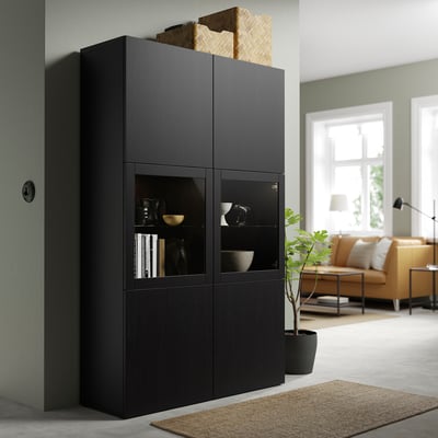 Unidade de almacenamento negro BESTÅ con portas de vidro, que alberga obxectos decorativos e libros. Está alto xunto a unha zona de estar cun sofá, lámpada e planta.