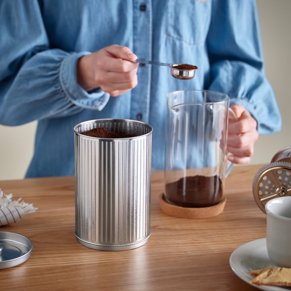 Unha persoa está a coller os pousos de café nun recipiente metálico sobre unha mesa, cunha prensa francesa de cristal e unha cunca preto.