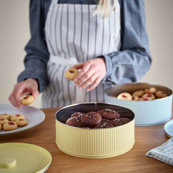 Persoa que sostén a galleta, rodeada de recipientes de produtos de forno. Cuncas e pratos cheos de galletas sobre mesa de madeira.