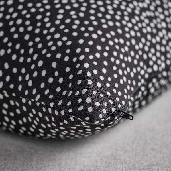 Cuberta de coxín negra con patrín branco punteado, detalle de cremalleira, colocada nunha superficie gris clara.