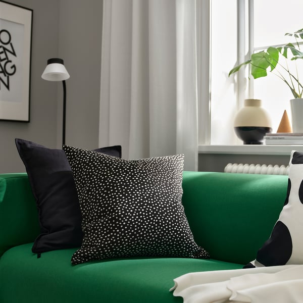 Elegante coxín verde BJÖRKGRÅMAL con patrón punteado nunha cama. Emparellado con coxín negro, contra parede gris con lámpada e planta. Decoración moderna.